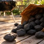 charcoal briquettes spilling out of an open paper bag onto a weathered wooden patio deck, a grill visible softly out of focus in the background, warm late-afternoon sunlight
