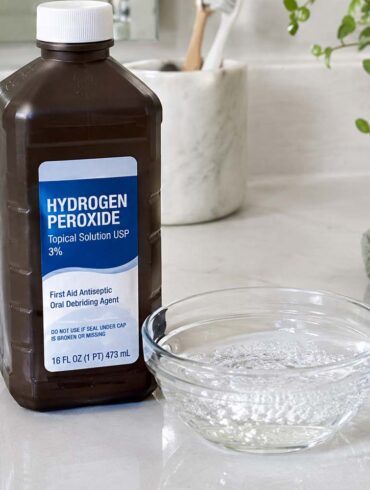 a brown hydrogen peroxide bottle on a white bathroom counter, a small stream of clear liquid fizzing gently in a glass dish beside it