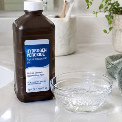 a brown hydrogen peroxide bottle on a white bathroom counter, a small stream of clear liquid fizzing gently in a glass dish beside it