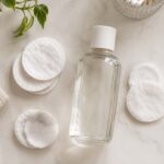 a clear glass bottle of nail polish remover with cotton pads scattered around it on a white bathroom vanity