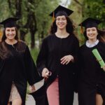 Three female graduates in academic robes, celebrating outdoors in a lush green environment.