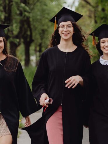 Three female graduates in academic robes, celebrating outdoors in a lush green environment.