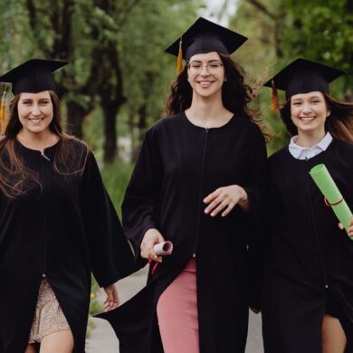 Three female graduates in academic robes, celebrating outdoors in a lush green environment.