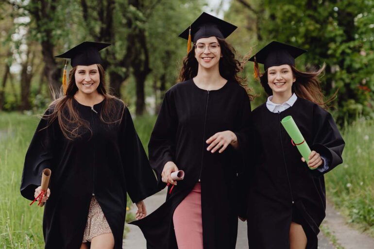 Three female graduates in academic robes, celebrating outdoors in a lush green environment.