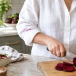 Close-up of a woman wearing a white button-down shirt while slicing deep purple beets on a wooden cutting board in a bright kitchen. A thin vivid magenta beet juice stain runs across the rolled-up sleeve of her shirt. Sliced beets, beet greens, a bowl of beet salad, a bottle of oil, salt, and a striped kitchen towel surround the cutting board on a light marble countertop, with natural window light and softly blurred kitchen details in the background.