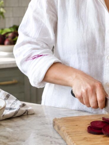 Close-up of a woman wearing a white button-down shirt while slicing deep purple beets on a wooden cutting board in a bright kitchen. A thin vivid magenta beet juice stain runs across the rolled-up sleeve of her shirt. Sliced beets, beet greens, a bowl of beet salad, a bottle of oil, salt, and a striped kitchen towel surround the cutting board on a light marble countertop, with natural window light and softly blurred kitchen details in the background.