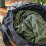 A close-up, photorealistic scene shows an open black gym bag resting on a rustic wooden bench, with a crumpled olive-green rain jacket partially pulled out of the bag. The jacket has scattered gray-green mildew spots concentrated across one shoulder and upper sleeve, with smaller spots visible on nearby folds, suggesting it was left damp inside the bag. A matte green water bottle is tucked along the side of the bag. In the softly blurred background, hiking shoes sit on the floor beside the bench, while a folded light-colored towel, tumbler, and leafy houseplant add natural details. Warm sunlight streams into the room, illuminating the wood and fabric and creating a realistic, lived-in post-hike setting.