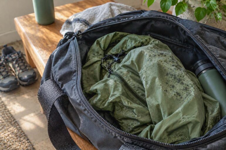A close-up, photorealistic scene shows an open black gym bag resting on a rustic wooden bench, with a crumpled olive-green rain jacket partially pulled out of the bag. The jacket has scattered gray-green mildew spots concentrated across one shoulder and upper sleeve, with smaller spots visible on nearby folds, suggesting it was left damp inside the bag. A matte green water bottle is tucked along the side of the bag. In the softly blurred background, hiking shoes sit on the floor beside the bench, while a folded light-colored towel, tumbler, and leafy houseplant add natural details. Warm sunlight streams into the room, illuminating the wood and fabric and creating a realistic, lived-in post-hike setting.
