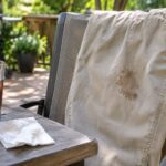A pair of light khaki pants draped over a patio chair has a small dried brown cola stain on the thigh. In the foreground, a glass of iced cola with a lime wedge sits beside a crumpled white napkin on a wooden patio table. A sunny backyard with greenery and a barbecue is softly blurred in the background.
