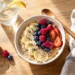 A bright overhead view of a bowl of creamy oatmeal topped with sliced banana, fresh strawberries, raspberries, blueberries, and chia seeds on a warm wooden table. A glass of water with a lemon wedge, small bowl of chia seeds, wooden spoon, and folded striped linen napkin surround the bowl, with soft morning sunlight casting gentle shadows across the breakfast scene.