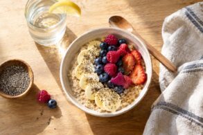 A bright overhead view of a bowl of creamy oatmeal topped with sliced banana, fresh strawberries, raspberries, blueberries, and chia seeds on a warm wooden table. A glass of water with a lemon wedge, small bowl of chia seeds, wooden spoon, and folded striped linen napkin surround the bowl, with soft morning sunlight casting gentle shadows across the breakfast scene.