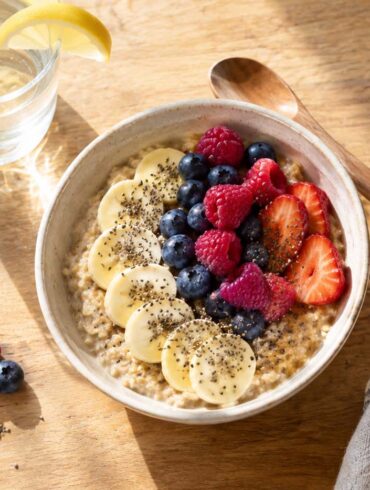 A bright overhead view of a bowl of creamy oatmeal topped with sliced banana, fresh strawberries, raspberries, blueberries, and chia seeds on a warm wooden table. A glass of water with a lemon wedge, small bowl of chia seeds, wooden spoon, and folded striped linen napkin surround the bowl, with soft morning sunlight casting gentle shadows across the breakfast scene.
