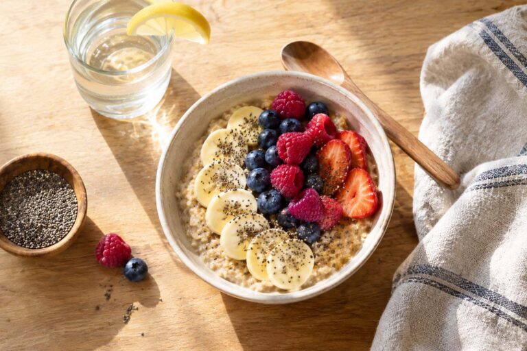 A bright overhead view of a bowl of creamy oatmeal topped with sliced banana, fresh strawberries, raspberries, blueberries, and chia seeds on a warm wooden table. A glass of water with a lemon wedge, small bowl of chia seeds, wooden spoon, and folded striped linen napkin surround the bowl, with soft morning sunlight casting gentle shadows across the breakfast scene.