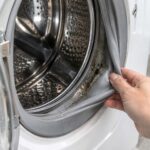 A hand pulls back the rubber gasket of an open front-loading washing machine, revealing dark mold and residue inside the damp seal.