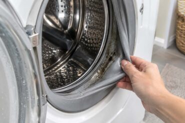 A hand pulls back the rubber gasket of an open front-loading washing machine, revealing dark mold and residue inside the damp seal.