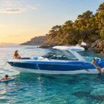 A sleek blue-and-white Formula powerboat anchored in crystal-clear turquoise water along a sunlit rocky coastline, with several people relaxing on and around the boat during golden hour.