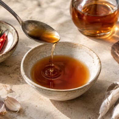 Fish sauce being poured from a spoon into a ceramic bowl, surrounded by red chilies, garlic cloves, lime halves, and a glass of amber fish sauce on a stone countertop.