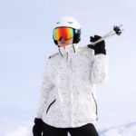 Skier wearing a white helmet, rainbow-mirrored ski goggles, and a white floral-print winter jacket, holding ski poles over one shoulder with a black-gloved hand, set against a pale blue sky.