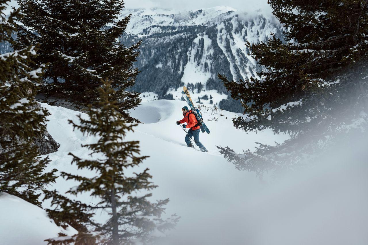 A skier in an orange jacket hikes uphill through deep snow with skis strapped to their backpack, framed by dark evergreen trees in the foreground and misty, snow-covered mountains in the background.