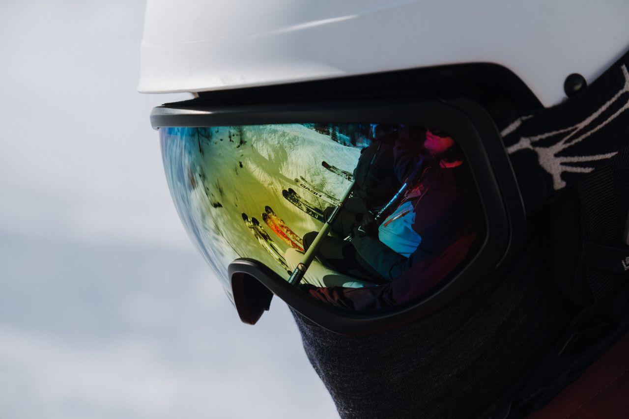 Close-up of a reflective ski goggle lens mounted on a white helmet, showing a mirrored reflection of a snowy ski slope with other skiers and their equipment visible in the glass.