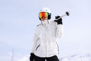 Skier wearing a white helmet, rainbow-mirrored ski goggles, and a white floral-print winter jacket, holding ski poles over one shoulder with a black-gloved hand, set against a pale blue sky.