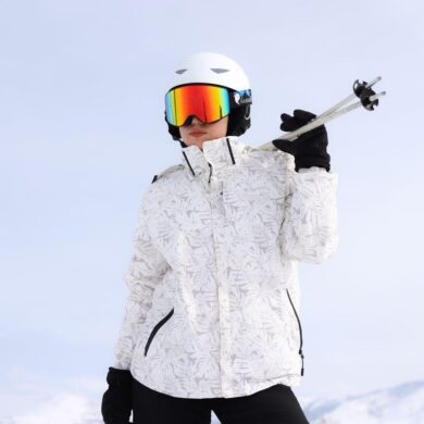 Skier wearing a white helmet, rainbow-mirrored ski goggles, and a white floral-print winter jacket, holding ski poles over one shoulder with a black-gloved hand, set against a pale blue sky.