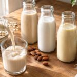 A close-up of three unlabeled glass bottles containing different types of plant-based milk, including beige oat milk, white almond milk, and pale golden milk, arranged on a warm wooden kitchen counter. One bottle is tilted and pouring milk into a clear glass, while rolled oats, whole almonds, shredded coconut, a whole coconut, fresh herbs, and a linen cloth surround the bottles in soft natural window light.