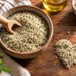 A rustic ceramic bowl filled with pale green and cream-colored hemp hearts sits on a richly textured wooden table, with a small wooden scoop resting in the seeds. A heart-shaped mound of hemp hearts is arranged beside the bowl, while a second small bowl of seeds, golden oil in a glass bottle, fresh green leaves, and a loosely draped striped linen cloth add natural detail around the scene. Warm sunlight highlights the texture of the hemp hearts and wood, creating an inviting, earthy food-photography composition.