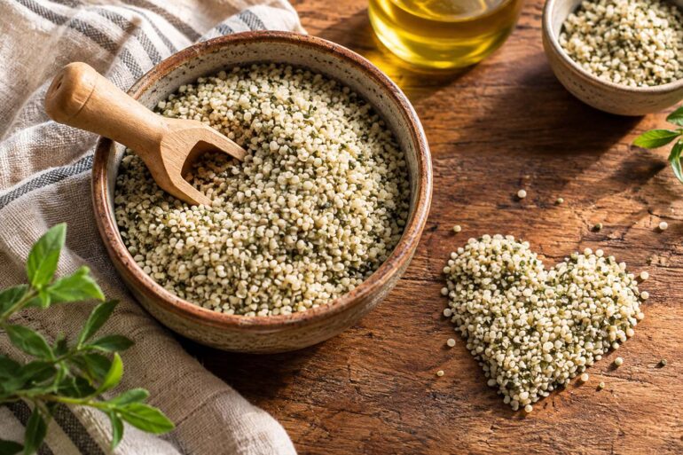 A rustic ceramic bowl filled with pale green and cream-colored hemp hearts sits on a richly textured wooden table, with a small wooden scoop resting in the seeds. A heart-shaped mound of hemp hearts is arranged beside the bowl, while a second small bowl of seeds, golden oil in a glass bottle, fresh green leaves, and a loosely draped striped linen cloth add natural detail around the scene. Warm sunlight highlights the texture of the hemp hearts and wood, creating an inviting, earthy food-photography composition.
