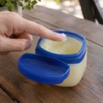 A close-up of a hand dipping a fingertip into an open jar of petroleum jelly with a blue lid on a wooden table. The jar contains translucent, pale yellow jelly, with the lid resting beside it. A folded white towel with blue stripes and green foliage are softly blurred in the background.