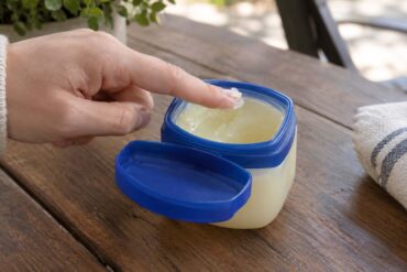 A close-up of a hand dipping a fingertip into an open jar of petroleum jelly with a blue lid on a wooden table. The jar contains translucent, pale yellow jelly, with the lid resting beside it. A folded white towel with blue stripes and green foliage are softly blurred in the background.