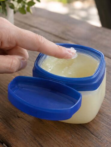 A close-up of a hand dipping a fingertip into an open jar of petroleum jelly with a blue lid on a wooden table. The jar contains translucent, pale yellow jelly, with the lid resting beside it. A folded white towel with blue stripes and green foliage are softly blurred in the background.