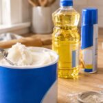 A close-up of an open blue shortening container on a wooden kitchen counter, with a spoon lifting a large scoop of white shortening from inside. In the softly blurred background are a bottle of cooking oil and an aerosol cooking spray can, positioned among baking ingredients including flour, a rolling pin, measuring dishes, and eggs.