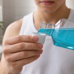 Close-up, bathroom scene showing a person pouring bright blue mouthwash from a clear bottle into a small white measuring cup. The person wears a white sleeveless shirt, with their hands, bottle, cup, and stream of mouthwash in sharp focus. A softly blurred bathroom vanity, folded white towel, green houseplant, and light-colored tile appear in the background.