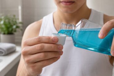 Close-up, bathroom scene showing a person pouring bright blue mouthwash from a clear bottle into a small white measuring cup. The person wears a white sleeveless shirt, with their hands, bottle, cup, and stream of mouthwash in sharp focus. A softly blurred bathroom vanity, folded white towel, green houseplant, and light-colored tile appear in the background.