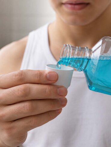 Close-up, bathroom scene showing a person pouring bright blue mouthwash from a clear bottle into a small white measuring cup. The person wears a white sleeveless shirt, with their hands, bottle, cup, and stream of mouthwash in sharp focus. A softly blurred bathroom vanity, folded white towel, green houseplant, and light-colored tile appear in the background.