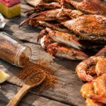 An overhead shot of a rustic wooden table set for a seafood feast. A pile of orange-red steamed blue crabs sits center and back, next to a mound of seasoned shrimp on the right. In the foreground, a glass jar has tipped over, spilling a heap of reddish-brown Old Bay seasoning across the wood, with a wooden spoon resting in the pile. Lemon wedges and crumpled paper towels are scattered around the edges, along with a white plate and the yellow-and-red Old Bay tin in the upper left.