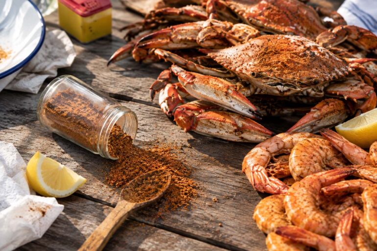 An overhead shot of a rustic wooden table set for a seafood feast. A pile of orange-red steamed blue crabs sits center and back, next to a mound of seasoned shrimp on the right. In the foreground, a glass jar has tipped over, spilling a heap of reddish-brown Old Bay seasoning across the wood, with a wooden spoon resting in the pile. Lemon wedges and crumpled paper towels are scattered around the edges, along with a white plate and the yellow-and-red Old Bay tin in the upper left.