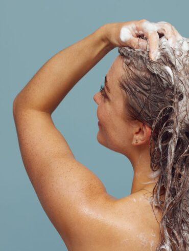 Woman with long hair washing it with shampoo, creating lather, in indoor setting