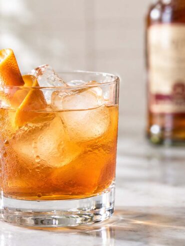 close-up of a SoCo amber-colored cocktail served over large ice cubes in a clear rocks glass on a pale marble countertop. The chilled glass is covered with natural condensation and garnished with a curled orange peel on a decorative cocktail pick. A Southern Comfort bottle sits softly blurred in the background, while natural daylight and a softly out-of-focus plant create a bright, warm kitchen-bar setting.