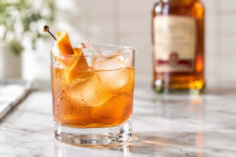 close-up of a SoCo amber-colored cocktail served over large ice cubes in a clear rocks glass on a pale marble countertop. The chilled glass is covered with natural condensation and garnished with a curled orange peel on a decorative cocktail pick. A Southern Comfort bottle sits softly blurred in the background, while natural daylight and a softly out-of-focus plant create a bright, warm kitchen-bar setting.