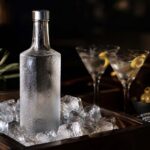 a frosted glass bottle of vodka standing on ice in a dark wood bar cart, condensation on the glass, two chilled cocktail glasses softly out of focus in the background