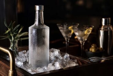 a frosted glass bottle of vodka standing on ice in a dark wood bar cart, condensation on the glass, two chilled cocktail glasses softly out of focus in the background