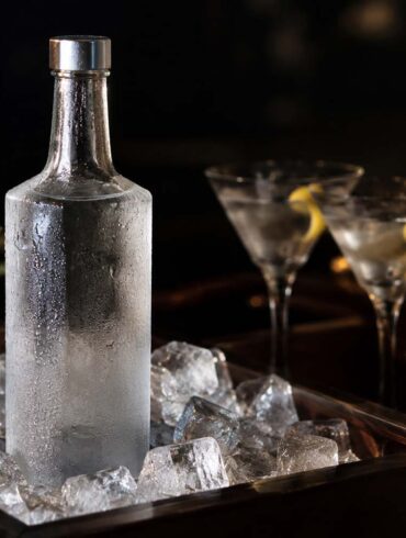 a frosted glass bottle of vodka standing on ice in a dark wood bar cart, condensation on the glass, two chilled cocktail glasses softly out of focus in the background