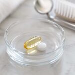 close-up photo of a small glass dish holding an omega-3 softgel capsule and a low-dose aspirin tablet, set against a soft neutral background with a subtle dental mirror or toothbrush blurred in the background
