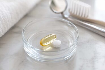close-up photo of a small glass dish holding an omega-3 softgel capsule and a low-dose aspirin tablet, set against a soft neutral background with a subtle dental mirror or toothbrush blurred in the background