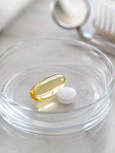 close-up photo of a small glass dish holding an omega-3 softgel capsule and a low-dose aspirin tablet, set against a soft neutral background with a subtle dental mirror or toothbrush blurred in the background