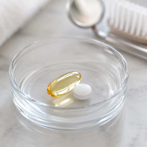 close-up photo of a small glass dish holding an omega-3 softgel capsule and a low-dose aspirin tablet, set against a soft neutral background with a subtle dental mirror or toothbrush blurred in the background