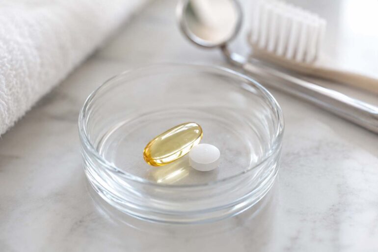close-up photo of a small glass dish holding an omega-3 softgel capsule and a low-dose aspirin tablet, set against a soft neutral background with a subtle dental mirror or toothbrush blurred in the background