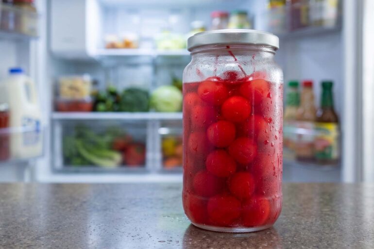 A chilled, clear glass jar of bright red maraschino cherries covered in condensation, sitting on a kitchen counter with an open, fully stocked refrigerator blurred in the background.